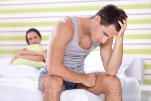 Uomo preoccupato seduto sul letto, donna sullo sfondo osserva.
